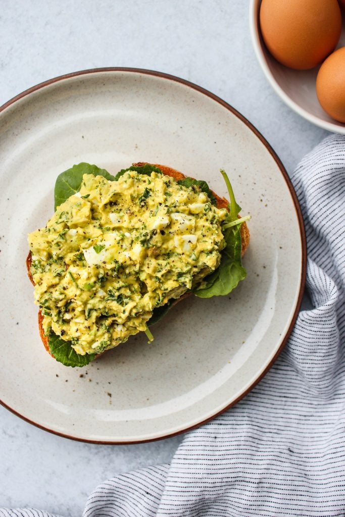 white plate with mashed avocado egg salad sandwich over a slice of toast