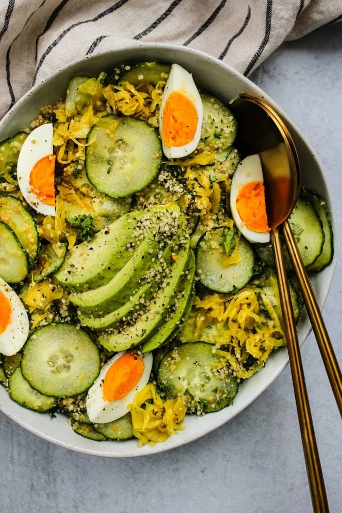 large white bowl with cucumber, avocado, quinoa, hard boiled eggs, sauerkraut salad and gold serving utensils