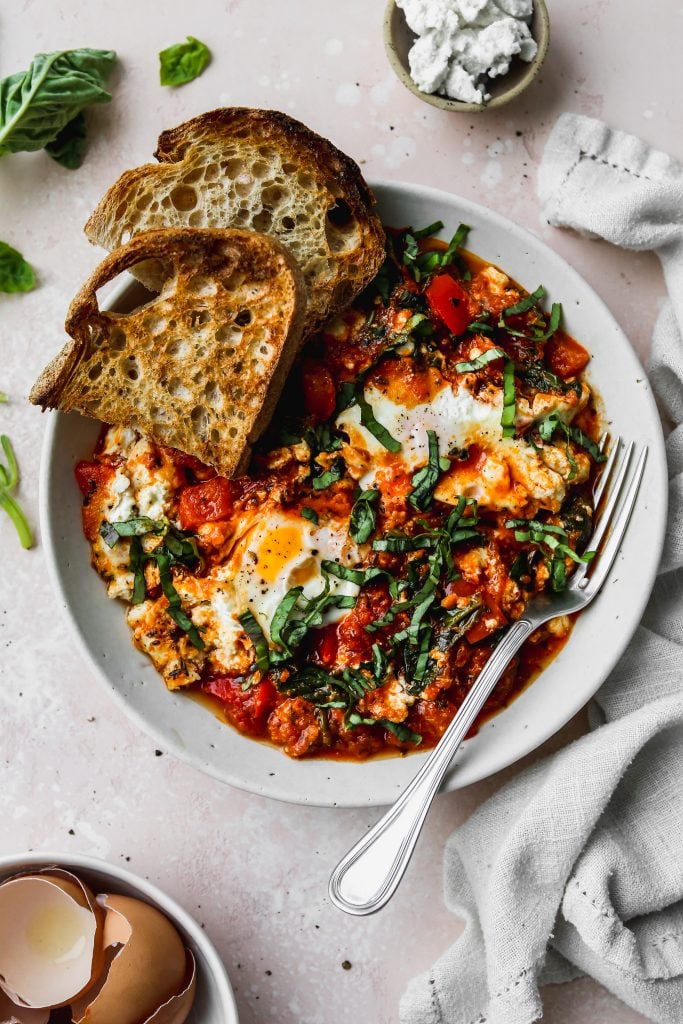 overhead photo of white bowl with a serving of egg shakshuka topped with basil and sourdough toast