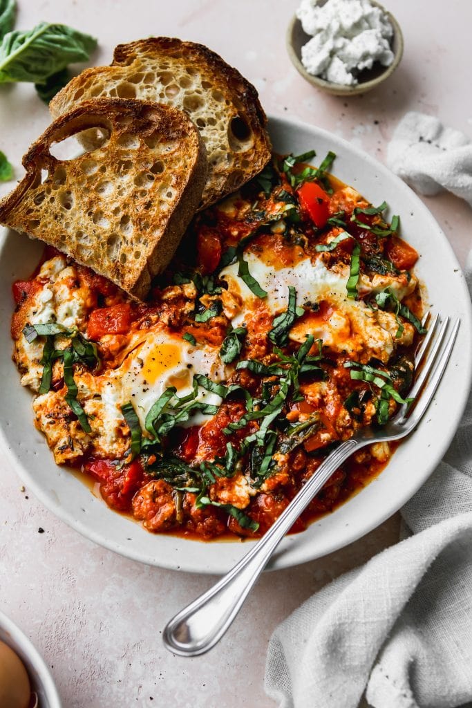 45 degree angle photo of white bowl with a serving of egg shakshuka topped with basil and sourdough toast