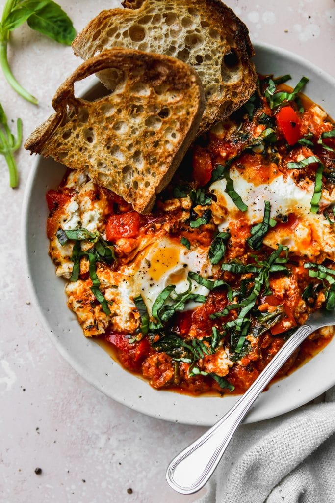 overhead photo of white bowl with a serving of egg shakshuka topped with basil and sourdough toast