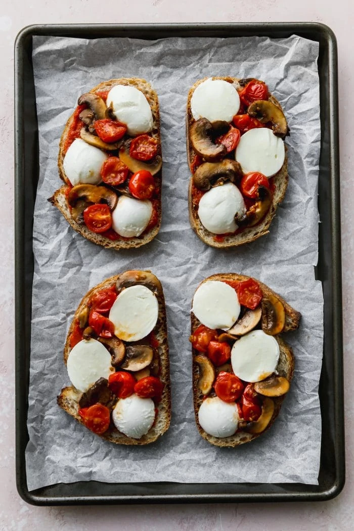 Overhead photo of a parchment-lined baking sheet topped with four slices of sourdough, bocconcini, tomatoes, and mushrooms.