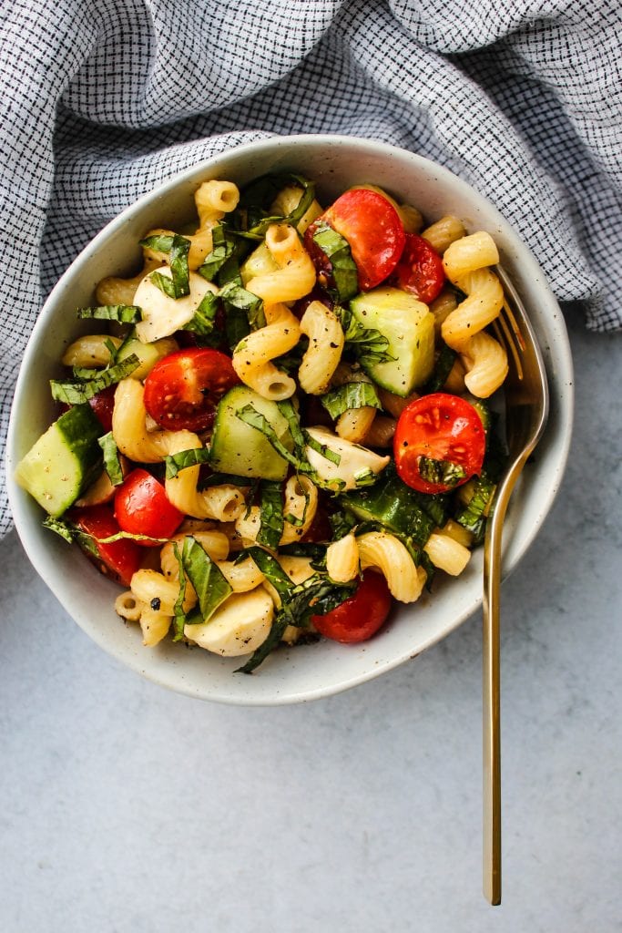 small bowl with cavatappi pasta, chopped cherry tomatoes, cucumbers, bocconcini, basil, and gold fork