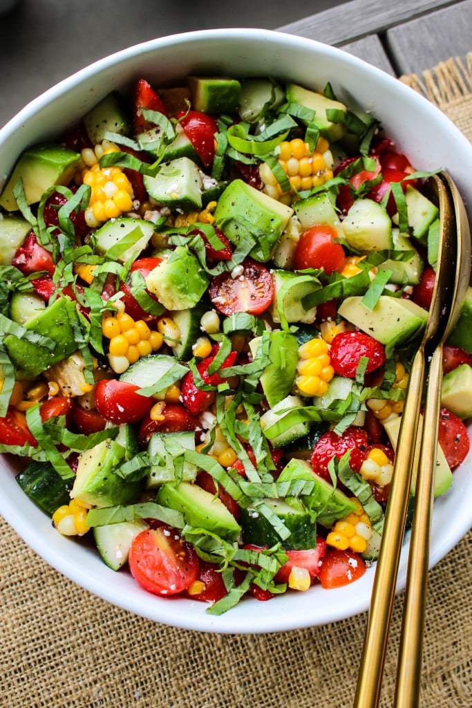 overhead shot of white bowl with corn, tomato, avocado, strawberry, basil, feta salad and gold serving spoons
