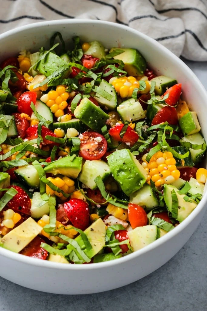 side angle shot of white bowl with corn, tomato, avocado, strawberry, basil, feta salad