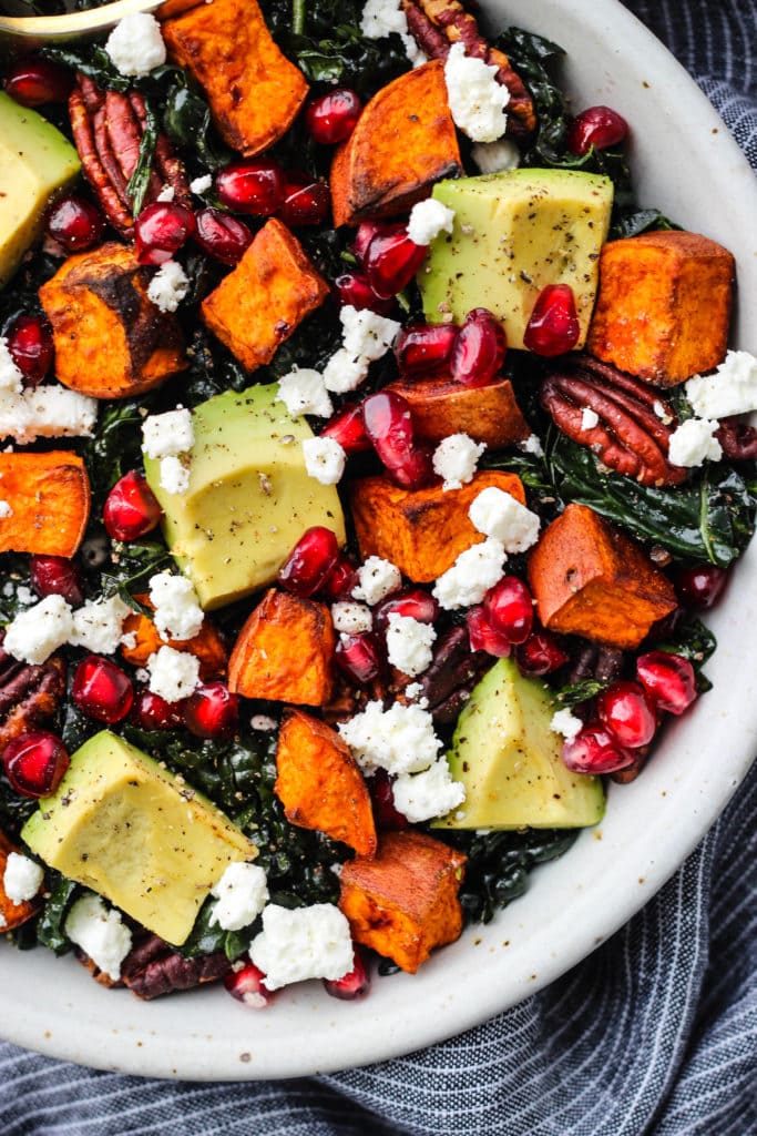 massaged kale salad with sweet potatoes, pomegranate, goat cheese, pecans, and avocado in white bowl