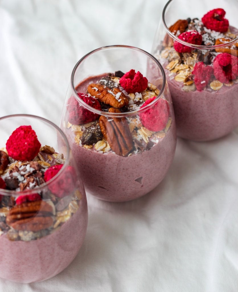 3 small glass cups of blended raspberry chia seed pudding topped with muesli, nuts, and seeds.
