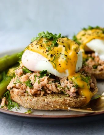 Whole wheat english muffin topped with canned salmon, a poached egg, hollandaise, and fresh parsley and dill on a plate.