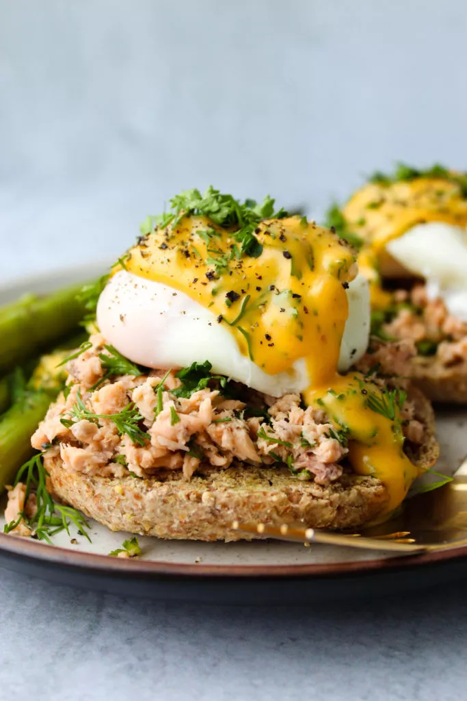 Whole wheat english muffin topped with canned salmon, a poached egg, hollandaise, and fresh parsley and dill on a plate.