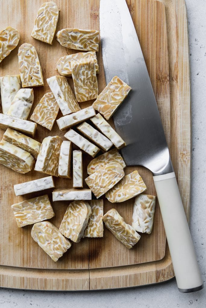 overhead photo of sliced tempeh on a wood cutting board with a chef's knife