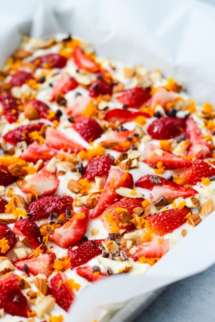 Baking sheet with frozen greek yogurt bark topped with chopped strawberries, orange zest, almonds, and cacao nibs
