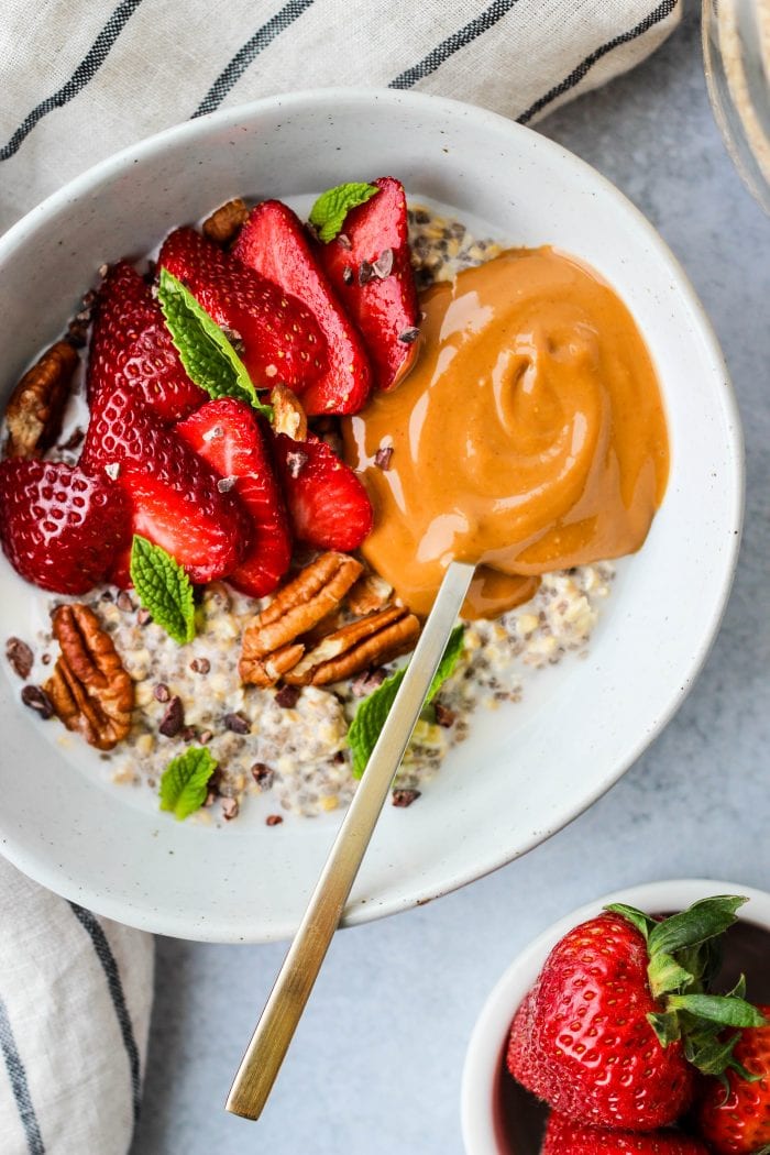 white bowl with steel cut oatmeal topped with sliced strawberries, mint leaves, pecans, and peanut butter