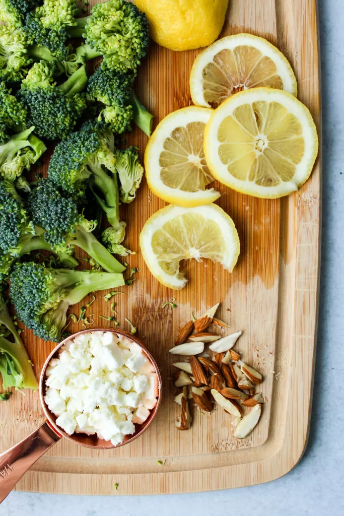wooden cutting board with broccoli florets, lemon slices, goat cheese, and chopped almonds