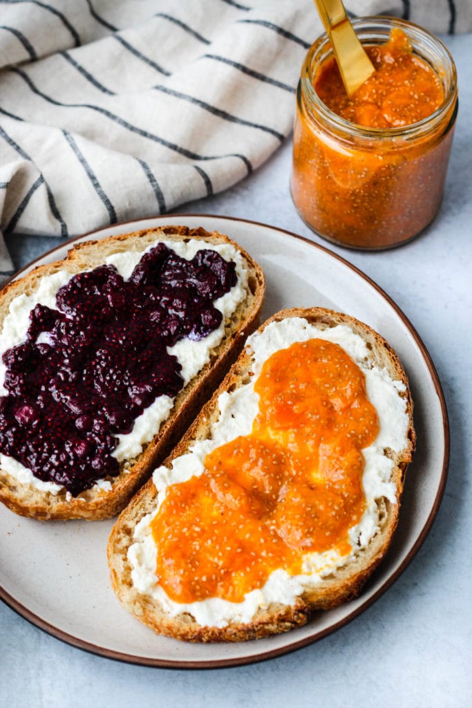 two slices of toast topped with ricotta and blueberry and apricot chia seed jam on white plate