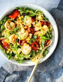 pasta salad with roasted red peppers, cherry tomatoes, corn, shrimp, and arugula in a white bowl, gold fork, and blue dish towel