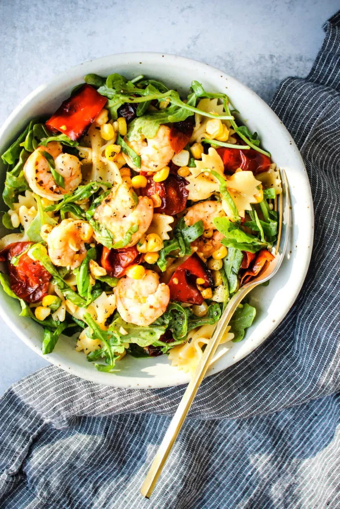 pasta salad with roasted red peppers, cherry tomatoes, corn, shrimp, and arugula in a white bowl, gold fork, and blue dish towel