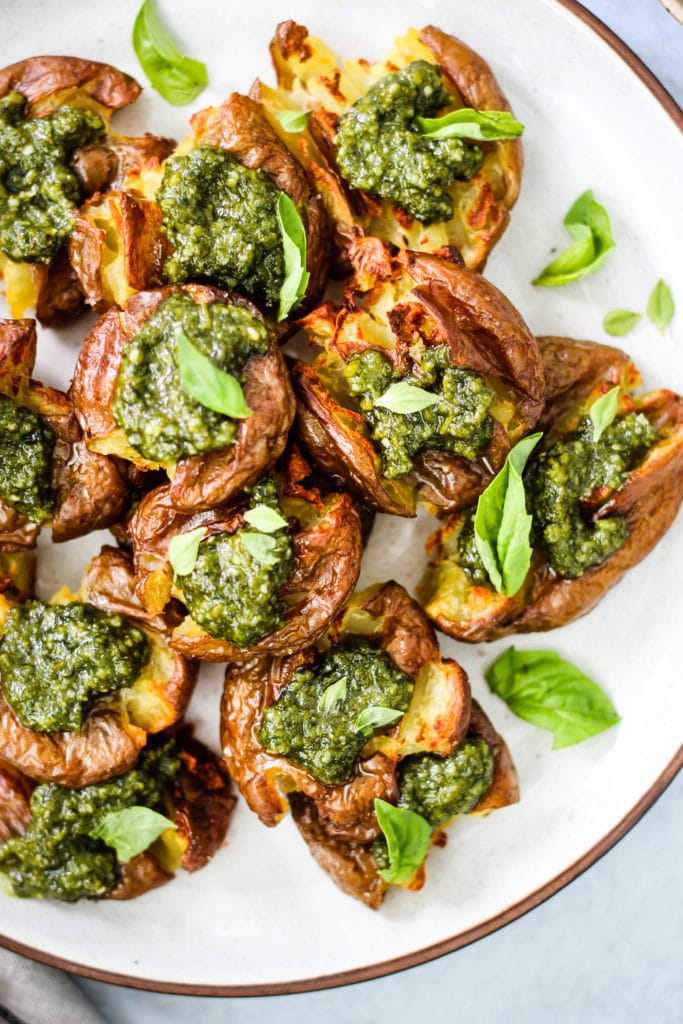 Overhead closeup photo of crispy red smashed potatoes topped with green pesto and basil leaves on white plate.