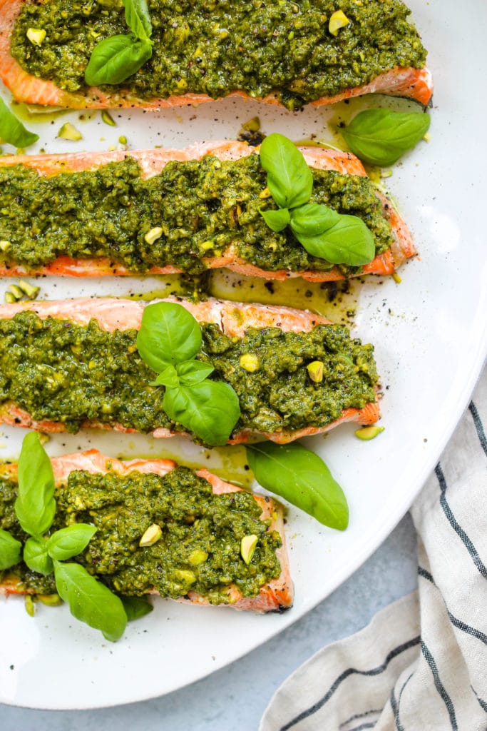 Overhead photo of 4 pieces of baked salmon topped with pistachio pesto and basil leaves on a large white serving platter.