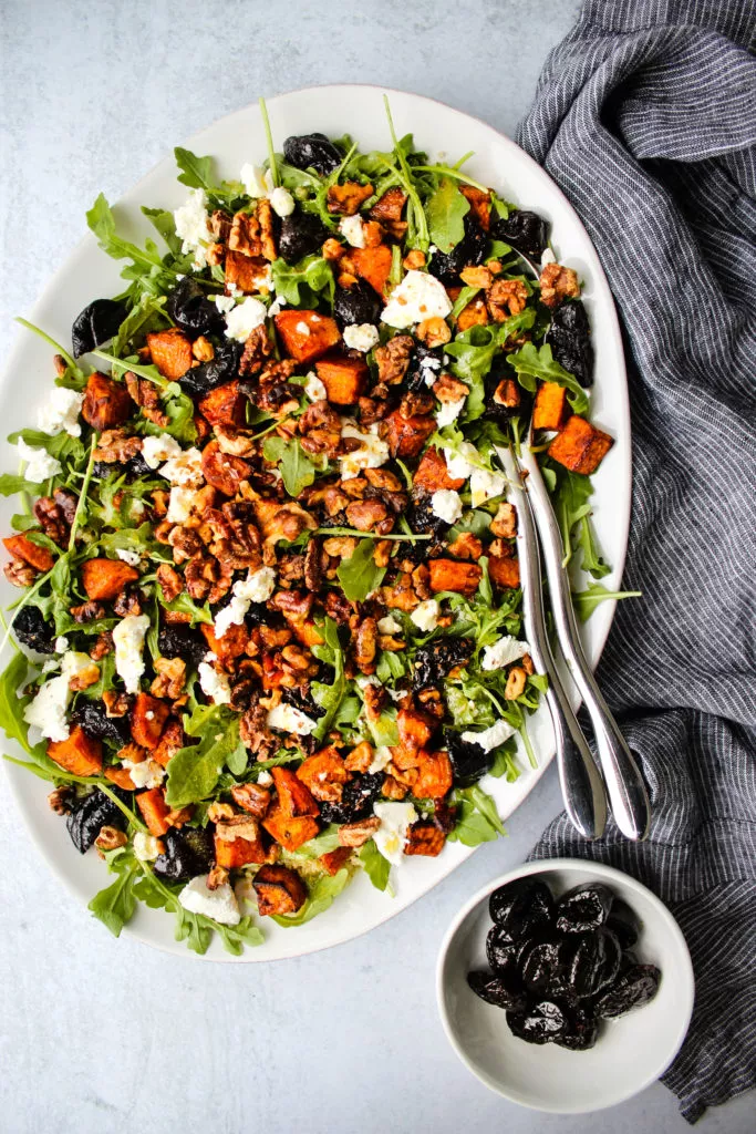 warm prune salad on white plate with arugula, sweet potatoes, goat cheese, and toasted walnuts