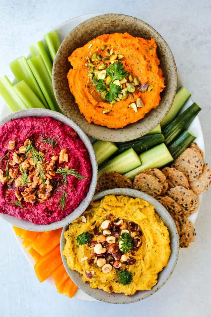three roasted vegetable dips in small bowls on serving platter with raw vegetables