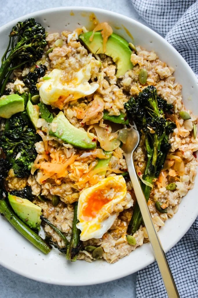 white bowl of oats mixed with kimchi, poached egg, broccolini, avocado slices