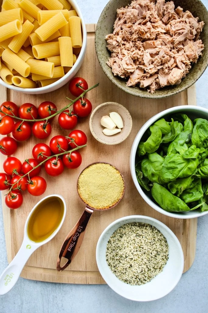wood cutting board with bowls of pasta, canned tuna, basil, hemp seeds, nutritional yeast, garlic, olive oil, and vine of cherry tomatoes