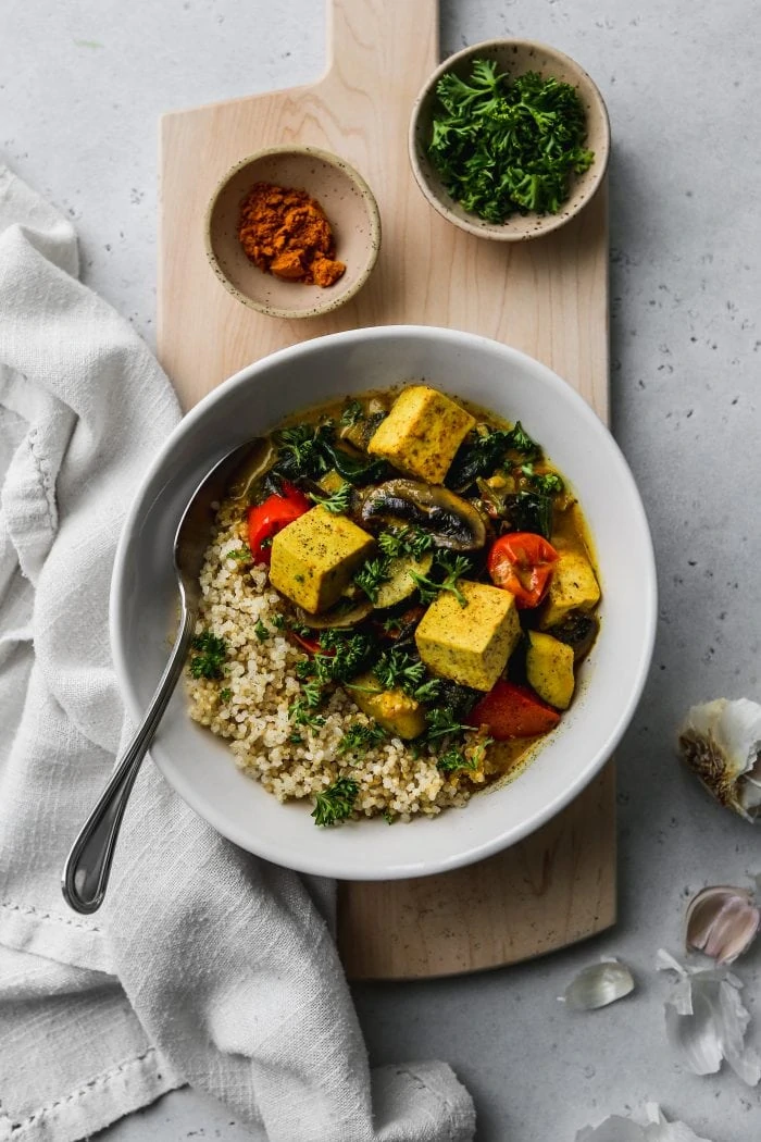 Overhead photo of a white bowl filled with quinoa, tofu, vegetables, and coconut curry on a wooden cutting board.