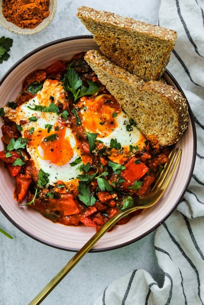 overhead shot of coconut curry shakshuka with two runny eggs in a pink bowl with two slices of toast and striped dishcloth
