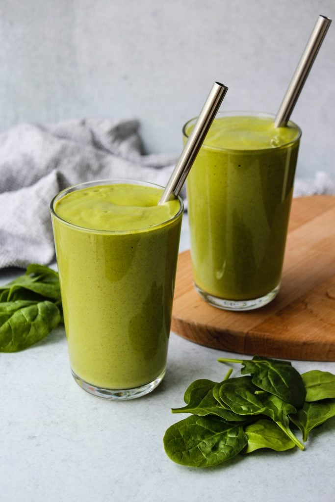 side view of two glasses of green smoothies with metal straws and baby spinach