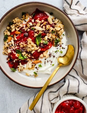 overhead shot of white bowl with greek yogurt, roasted strawberries, granola, mint, gold spoon and dishtowel