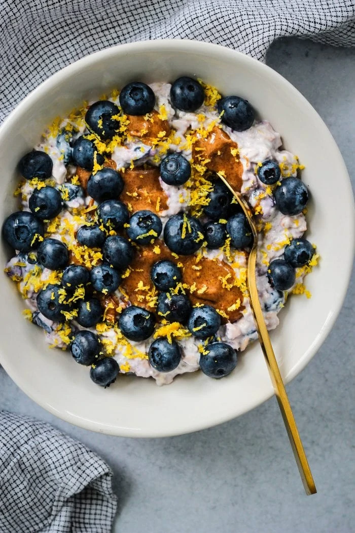 overhead shot of white bowl with overnight oats, blueberries, lemon zest, almond butter and gold spoon with blue towel