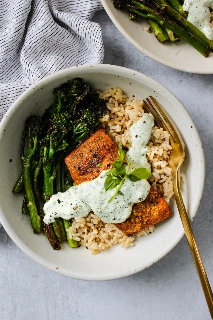overhead shot of white bowl with broccolini, brown rice, turmeric salmon, parsley yogurt sauce