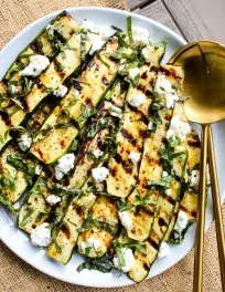 light blue plate with grilled zucchini slices topped with crumbled goat cheese, basil and honey and golden serving spoons