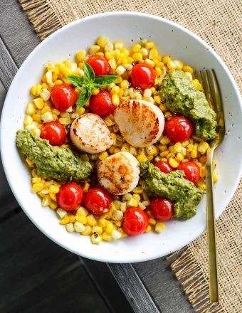 overhead shot of white bowl with bed of corn and cherry tomatoes topped with seared scallops and pesto