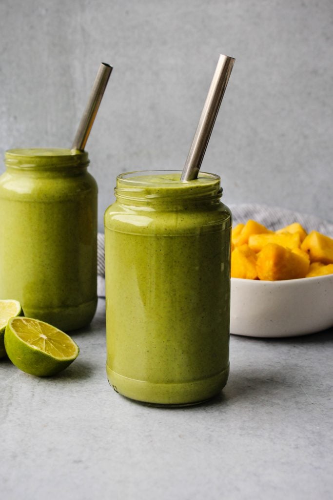 two glass jars filled with green smoothie and metal straws, lime and pineapple in the background