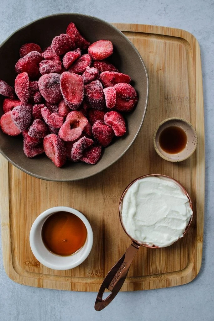Overhead photo of a wood cutting board with bowls of frozen strawberries, yogurt, maple syrup, and vanilla on top.