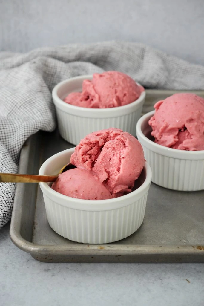 Side angle photo of three white ramekins on top of a baking sheet with scoops of homemade strawberry frozen yogurt inside.