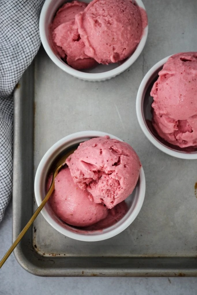 Overhead shot of baking sheet topped with three white ramekins holding scoops of homemade strawberry frozen yogurt inside.