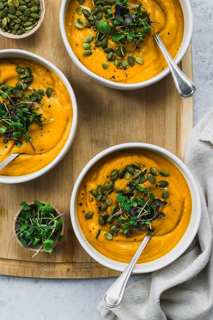 Overhead shot of three white bowls filled with butternut squash soup and topped with microgreens and pumpkin seeds.