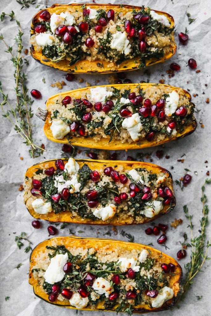 Overhead photo of 4 halves of delicata squash stuffed with kale and quinoa and topped with goat cheese, pomegranates, and thyme on white parchment paper.