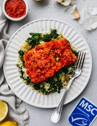 overhead photo of baked harissa salmon over lemon kale quinoa on a white plate. Ingredients garnish the outside of the plate, along with a MSC blue label