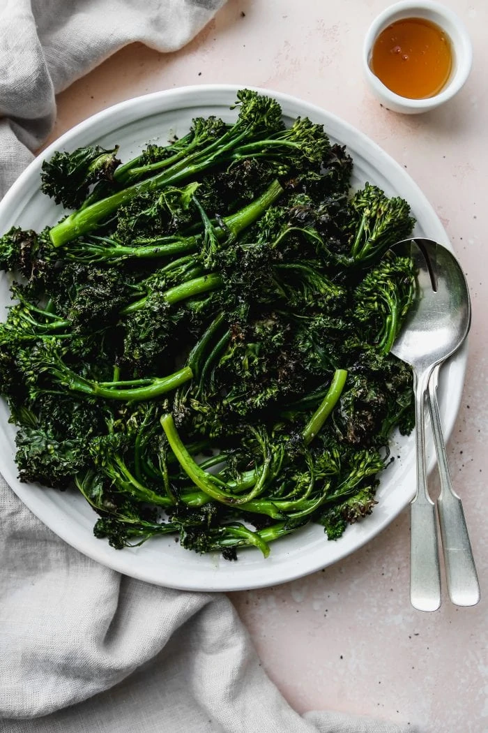 overhead photo of roasted broccolini and kale on a white serving platter with small bowl of honey and silver serving utensils