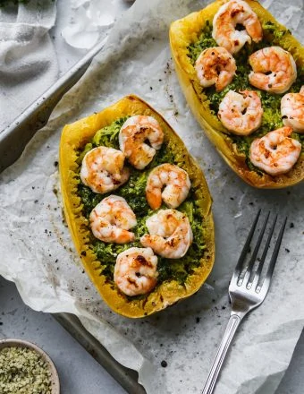 overhead photo of two halves of roasted spaghetti squash topped with pesto and shrimp on a parchment-linked baking sheet