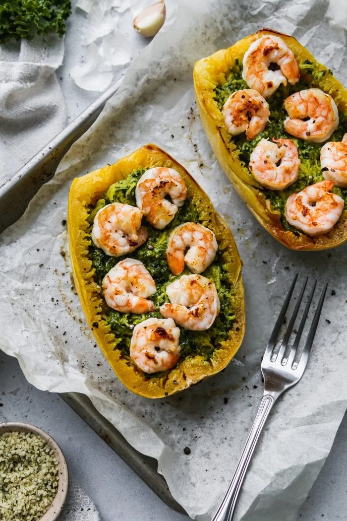 overhead photo of two halves of roasted spaghetti squash topped with pesto and shrimp on a parchment-linked baking sheet
