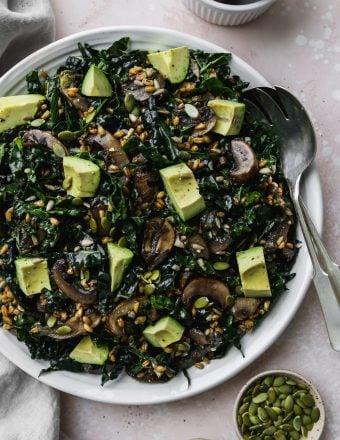 overhead photo of large white serving plate topped with warm mushroom, kale, and farro salad with avocado chunks on top
