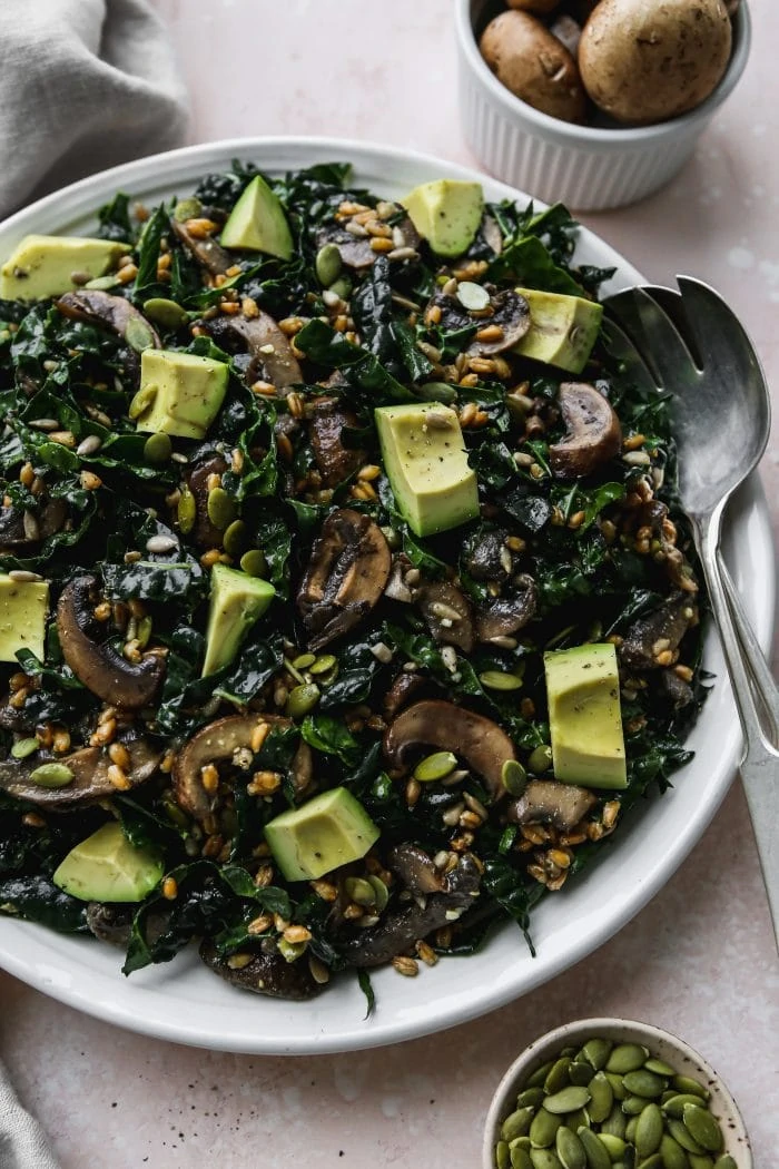 45 degree angle photo of large white serving plate topped with warm mushroom, kale, and farro salad with avocado chunks on top
