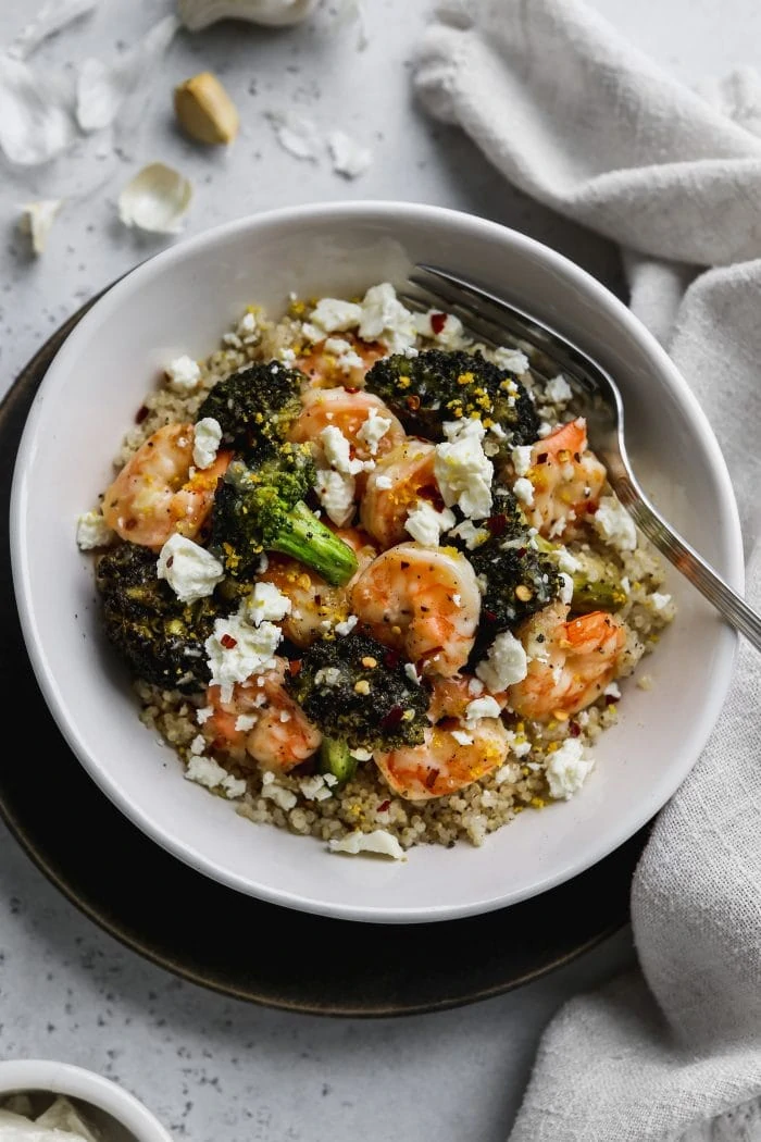 45 degree angle photo of white bowl filled with quinoa, sautéed shrimp, broccoli, feta, and lemon zest.
