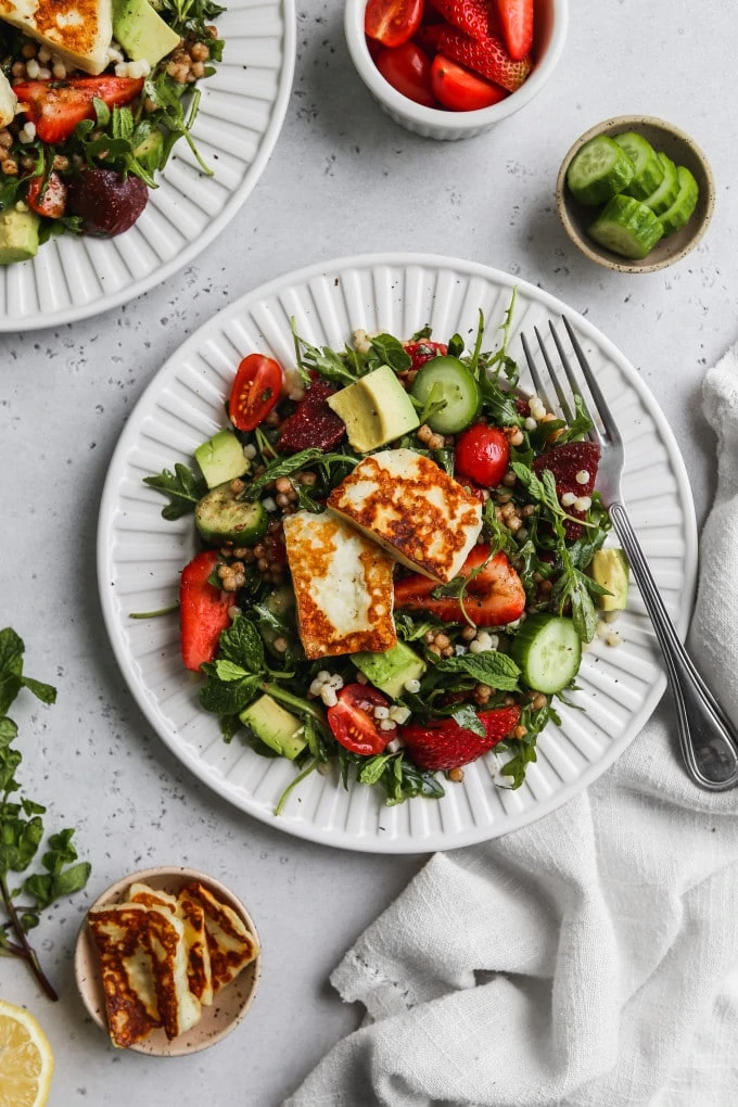Overhead photo of a white plate topped with an arugula, halloumi, couscous, and vegetable salad.