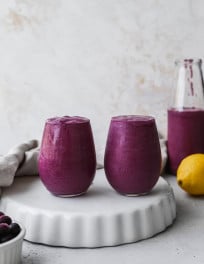 Straight on photo of two small glasses filled with blueberry lemon avocado smoothie on a white stand.