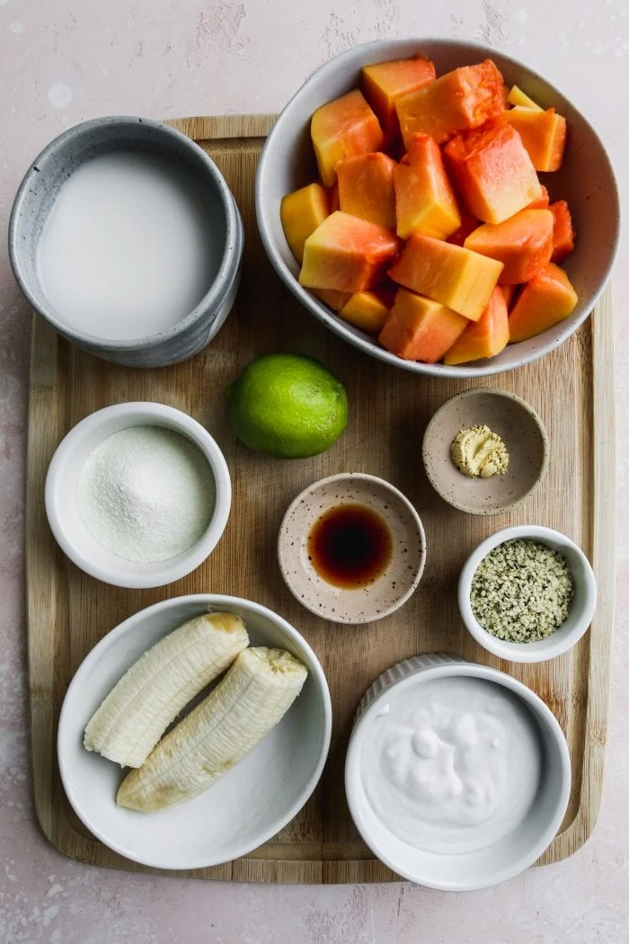 Overhead photo of a wooden cutting board with several small bowls filled with smoothie ingredients on top.